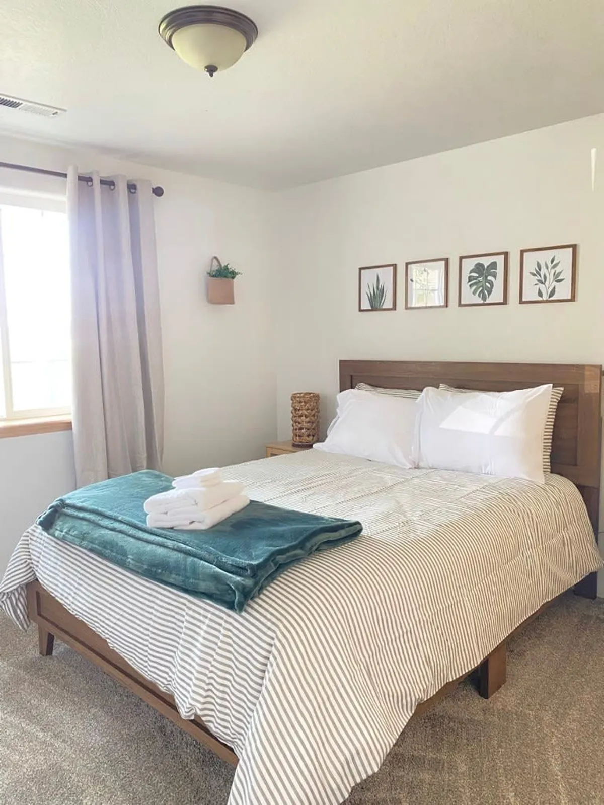 A second bedroom featuring a comfortable bed with striped bedding, framed botanical prints, a green plant on a shelf, and neatly folded towels on the bed. The overall atmosphere is calm, fresh, and inviting.