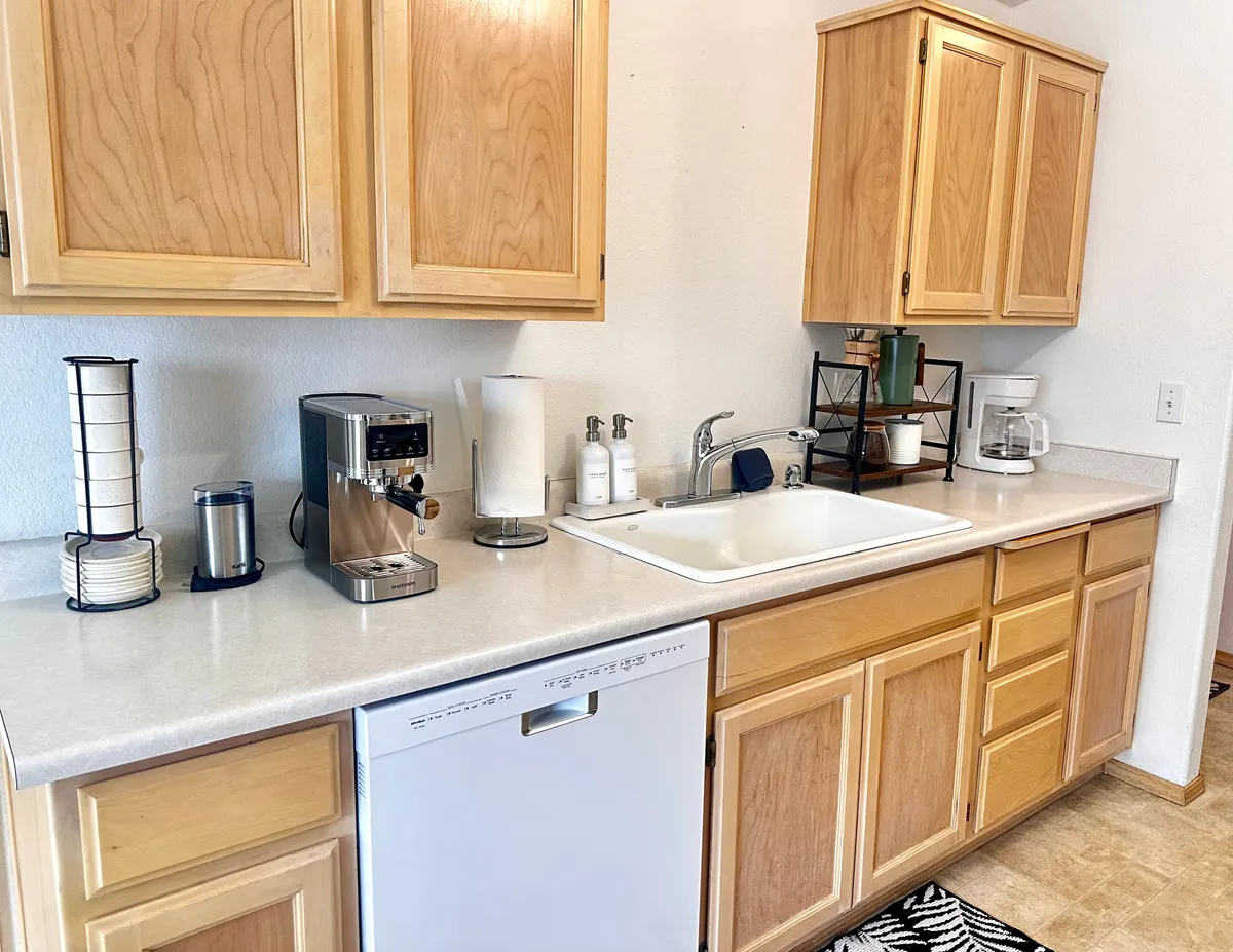 This image captures a welcoming kitchen space featuring a coffee maker, dishwasher, sink, and kitchen cabinets. The countertop has ample space for prepping meals, and the overall atmosphere is warm and inviting, suggesting a space where cooking and entertaining are everyday joys.