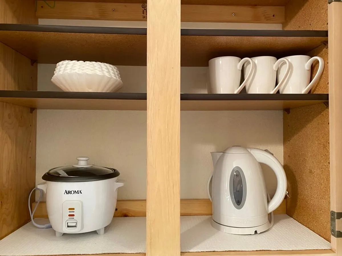 A tidy and organized kitchen counter with a white rice cooker, kettle, ceramic plates, and mugs, evoking the feeling of preparing and sharing warm meals or hot beverages with friends after a day out. The setup is welcoming and practical.