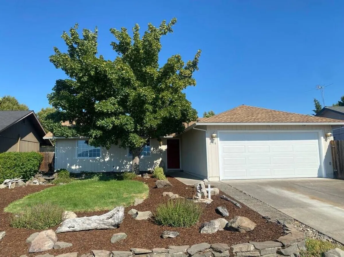 This exterior image captures a spacious green lawn accented by a large shade tree, colorful flower beds, and a well-maintained driveway. The scene suggests an inviting outdoor space perfect for family gatherings, barbecues, gardening, and playful moments with pets.