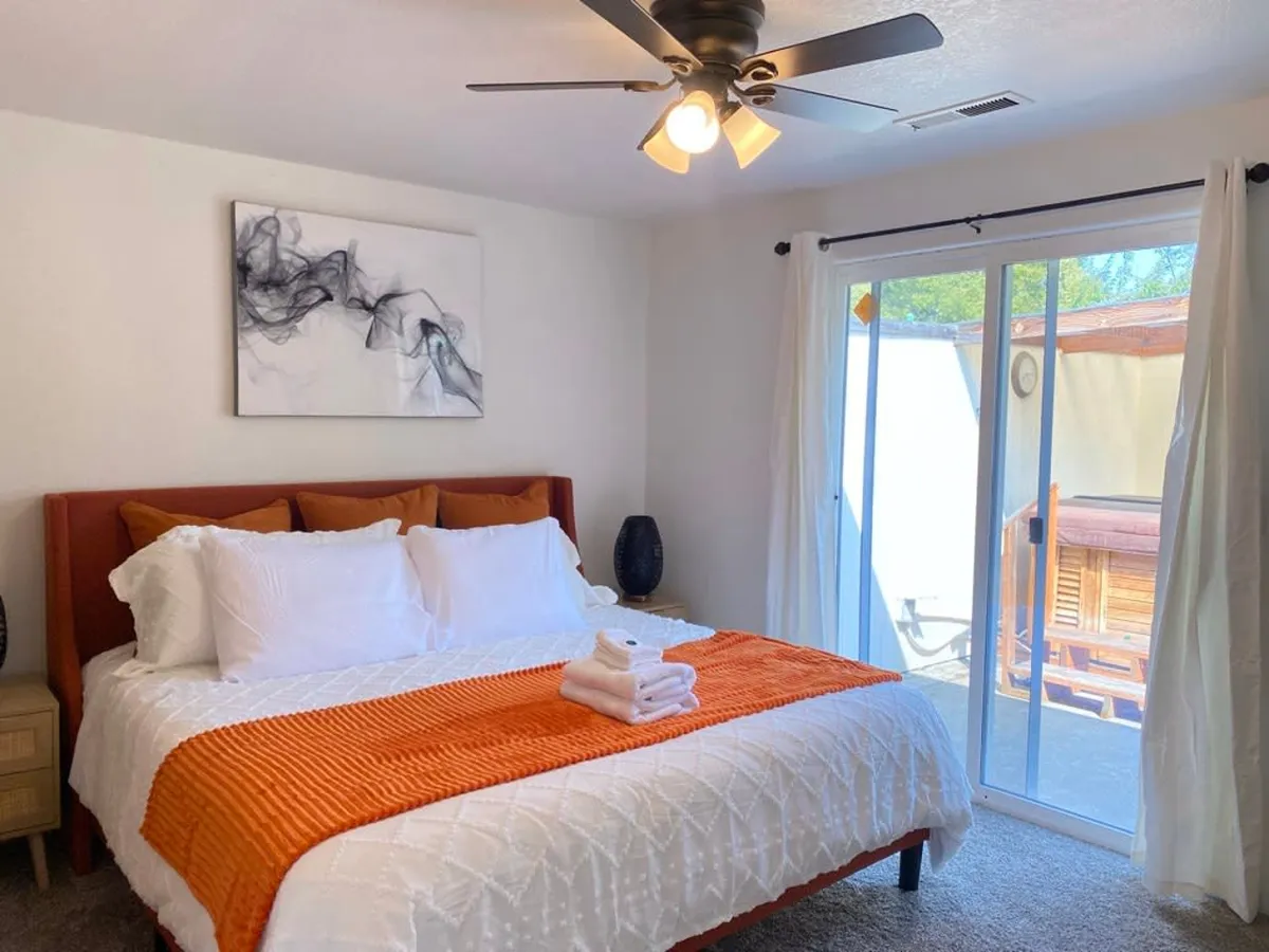 This image shows a serene bedroom with a plush bed adorned with an orange accent blanket, abstract wall art, and a sliding glass door that leads directly to outdoor space, likely the covered spa area. Natural light fills the room, emphasizing comfort and relaxation.