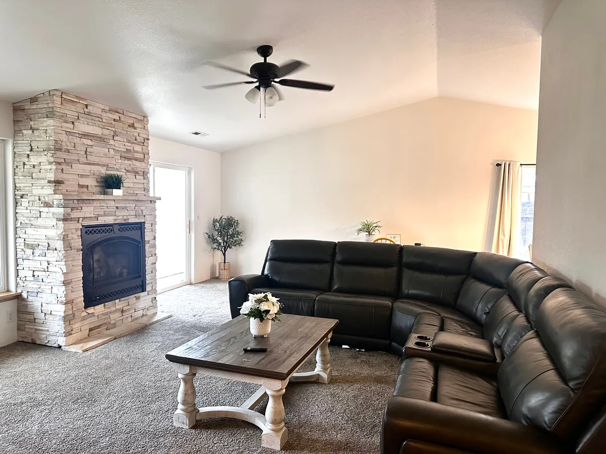 A spacious living area featuring a leather sectional couch facing a stone fireplace, with a wooden coffee table topped with fresh flowers. The room is filled with natural light and designed for both cozy gatherings and festive celebrations.
