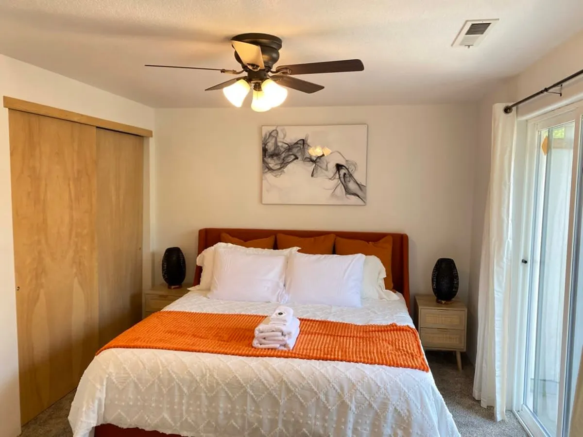 A cozy master bedroom with a plush bed, artwork, and nightstands. The room exudes comfort and relaxation, with an orange throw blanket adding a pop of color. Ample natural light hints at access to an adjoining space, suggesting the transition to the spa area described in the section.
